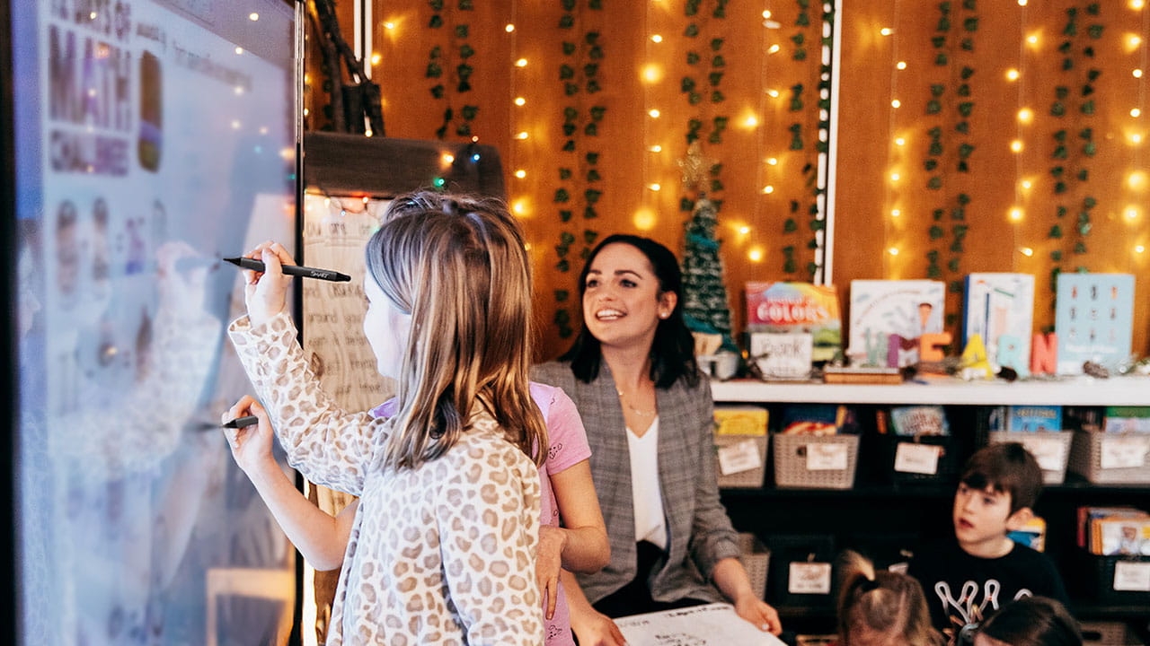 Students collaborating on a SMART Board in a decorated classroom with teacher supervision.